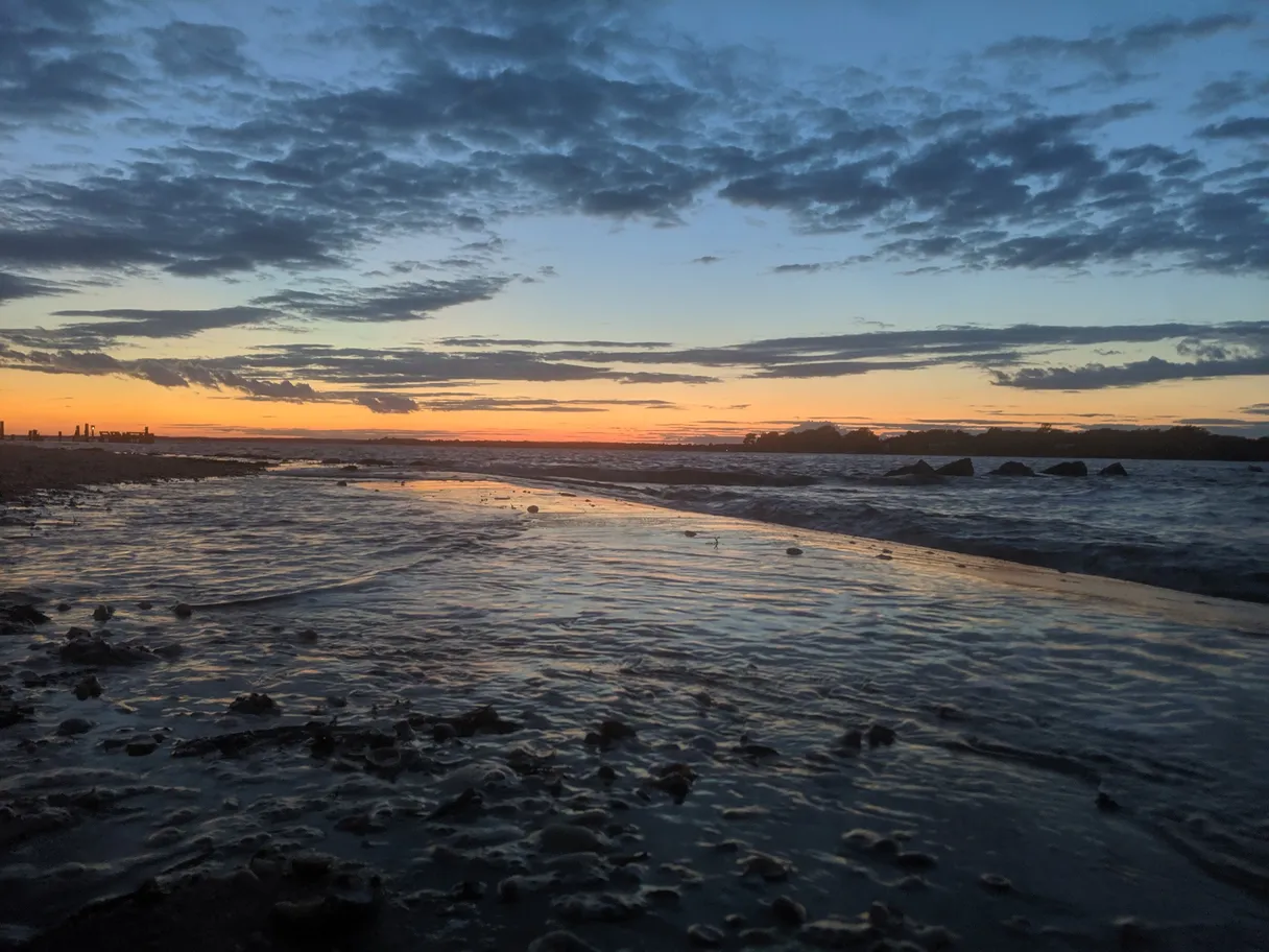 Sunset over the bay at Long Beach in Noyack, NY: blue clouds above a thin orange horizon, with shallow water washing over a rocky beach in the foreground